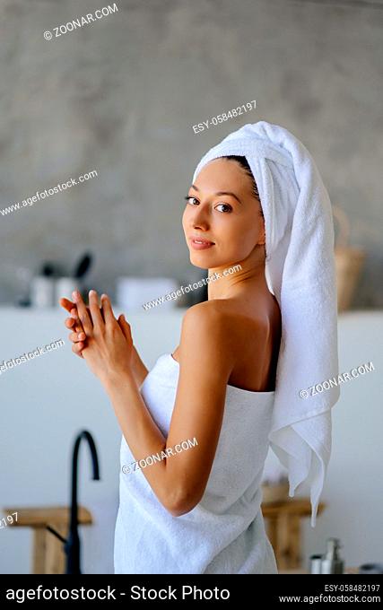 Relaxed young Caucasian female model in white towel, feels refreshed after taking shower, has healthy clean soft skin, poses in cozy bathroom