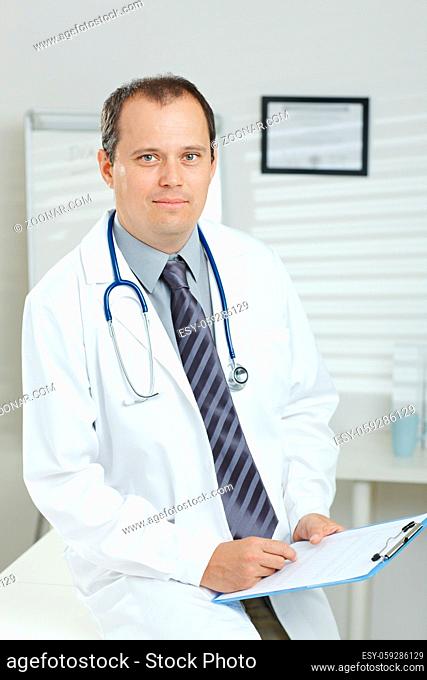 Medical office - middle-aged male doctor writing on clipboard, looking at camera, smiling