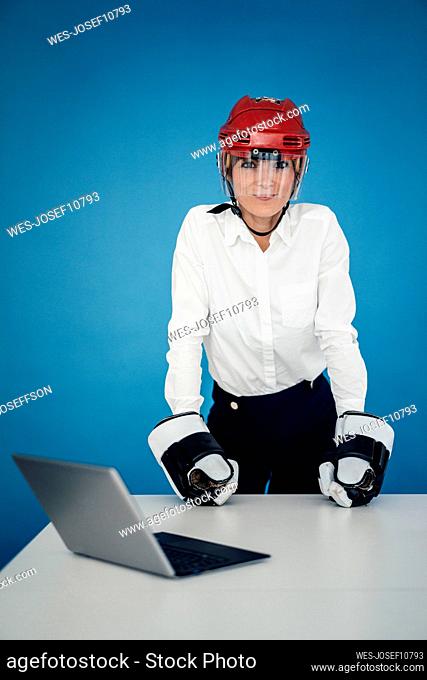 Businesswoman wearing ice hockey helmet and gloves at table in front of blue wall