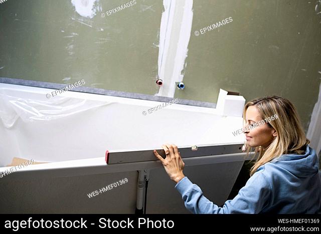 Blond woman measuring bathtub with spirit level in loft apartment