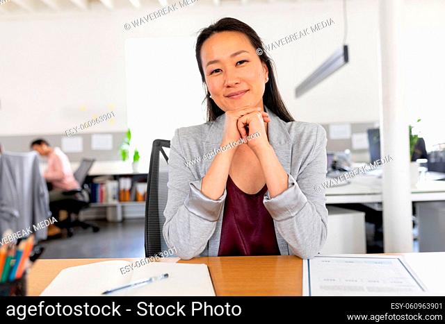 Portrait of confident asian creative businesswoman sitting with hands on chin at desk in office