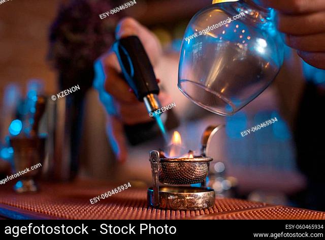 Closeup of male hands using professional burner to prepare drink with glass and strainer on bar counter