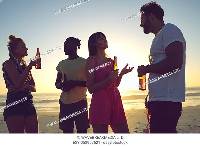 Side view of group of diverse friends having beer and interacting with each at beach during sunset