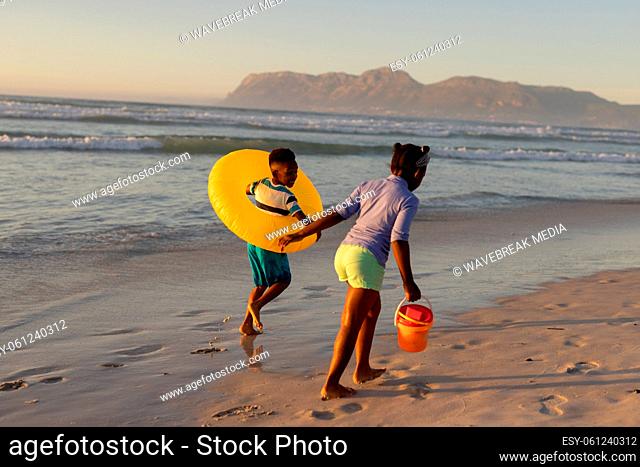 Playful african american boy with inflatable ring and girl with pail running at beach during sunset