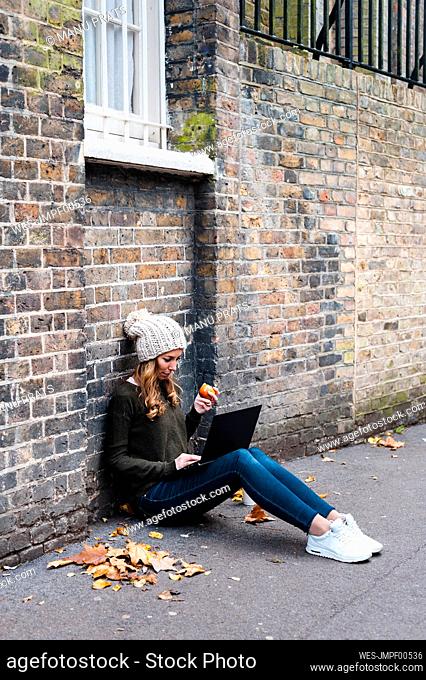 Woman using laptop while eating apple against brick wall on footpath