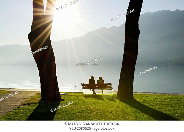 Couple sitting on a bench on the lakefront maggiore with brissago islands and mountain with sunbeam on a misty lake in ascona ticino switzerland