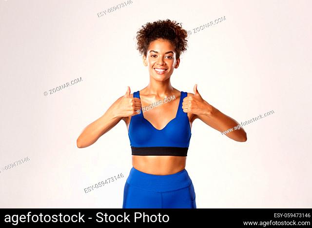 Waist-up of beautiful african-american fitness woman, wearing blue sport outfit, showing thumbs-up in approval, recommend gear for workout, white background