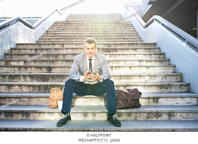 Confident businessman in the city sitting on stairs using cell phone