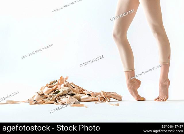 Pile of beige pointe shoes on the light background in the studio. Next to it there are ballerina's legs on the toes in beige dance wear. Closeup