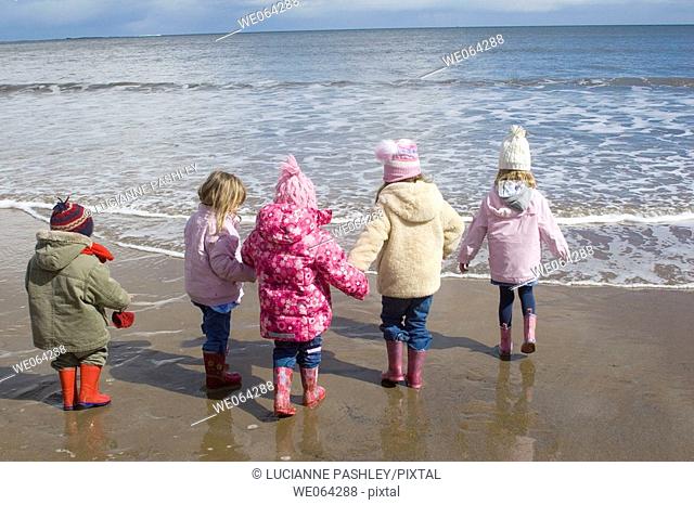 four girls and a boy standing in a line at the shore, holding hands, about to paddle in wellies