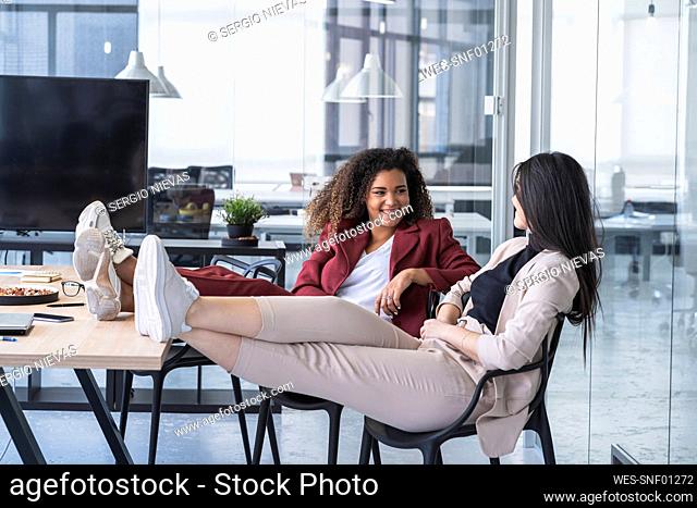 Smiling businesswomen discussing while sitting with feet up on chair at coworking office