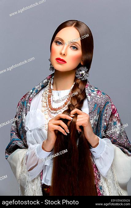 Beautiful young russian woman with very long braid in traditional shawl. Red lips. Over grey background