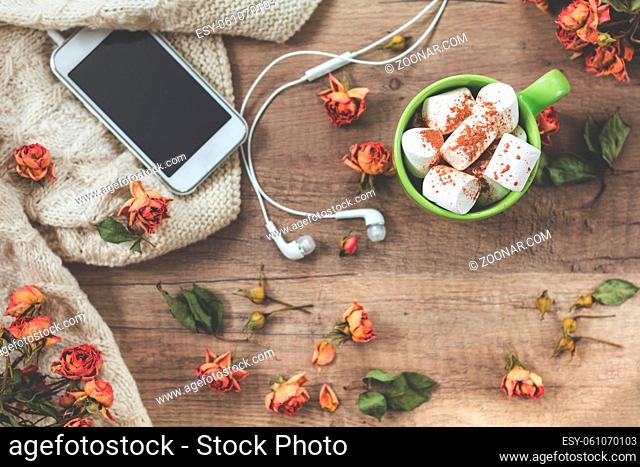 Cup of coffee with marshmallow, white knitting wool, dried roses flowers, mobile phone and headphones. Beautiful romantic female?s background