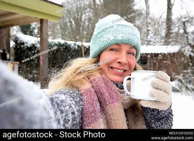 Smiling mature woman holding coffee cup while standing at backyard during snow