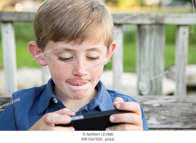 A young boy using a hand held electronic tablet or game and concentrating, sticking his tongue out