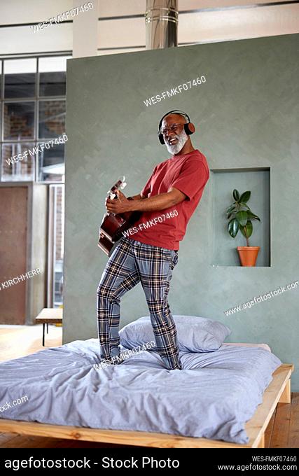 Mature man with headphones playing guitar on bed at home