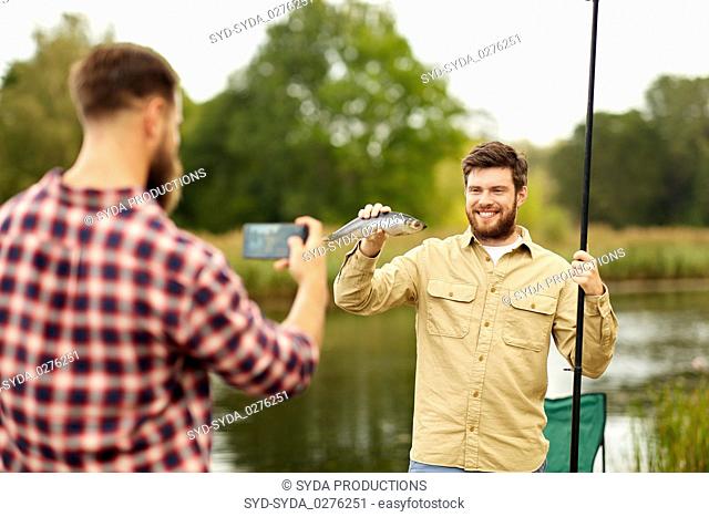 friend photographing fisherman with fish at lake