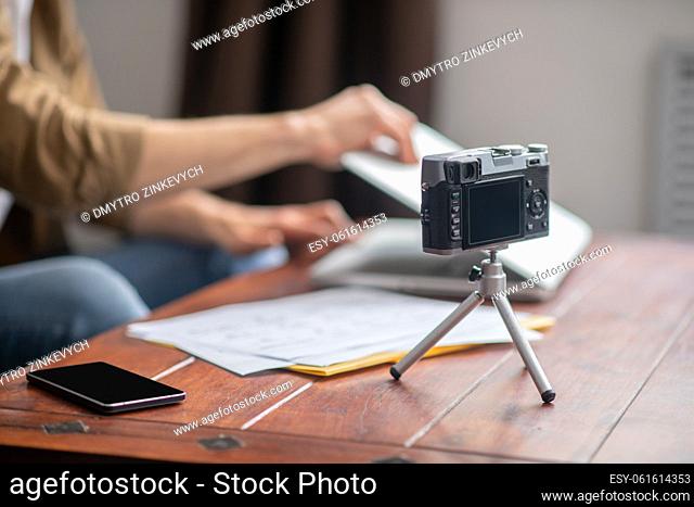 Device. Camera on stand and smartphone on table near pack of papers and hands opening laptop in room in daylight