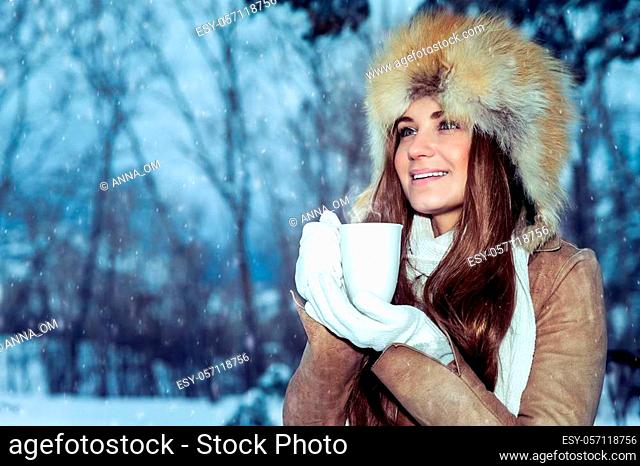 Portrait of a beautiful young girl wears a stylish hat standing in a snowy park and warming hands on a hot mug of tea, enjoying the beautiful winter view
