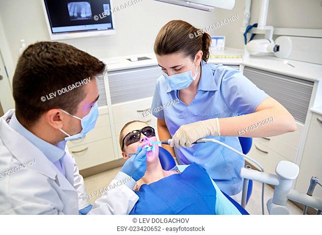people, medicine, stomatology and health care concept - male dentist and assistant with dental curing light and mirror treating female patient teeth at dental...