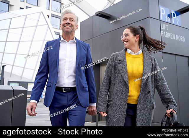 Smiling businesswoman and businessman talking while walking against elevator at bus stop in city