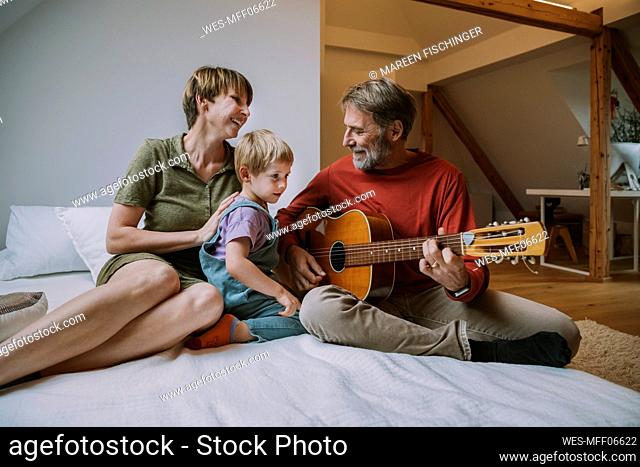 Father playing guitar while mother and son sitting on bed in bedroom at home