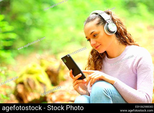 Relaxed woman wearing headphones listening music and wathing phone content in a forest