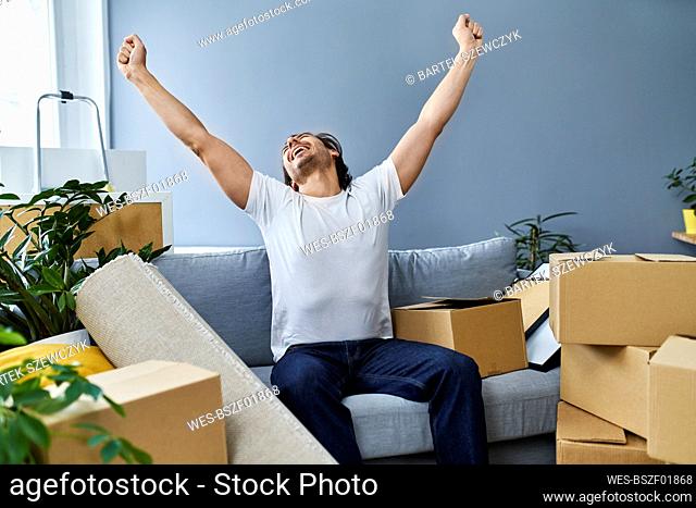Happy man with arms raised sitting on sofa amidst cardboard boxes at home