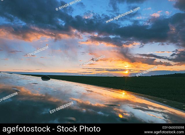 Sunset Sky With Setting Sun Reflected In Hood Of Car. Countryside Landscape Under Scenic Colorful Sky At Sunset Dawn Sunrise. Skyline. Springtime