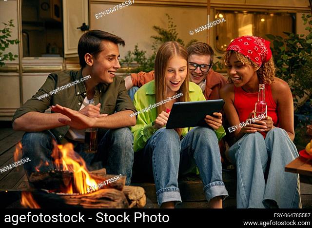 Happy friends spending evening nearby bonfire and campsite. Four millennial people watching video using mobile tablet smiling and laughing