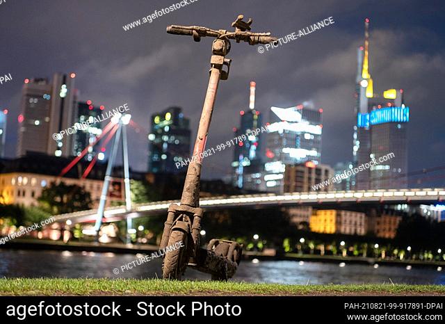 18 August 2021, Hessen, Frankfurt/Main: A muddy e-scooter stands on the banks of the Main River in front of the skyline. (to dpa ""Great demand for e-scooters -...