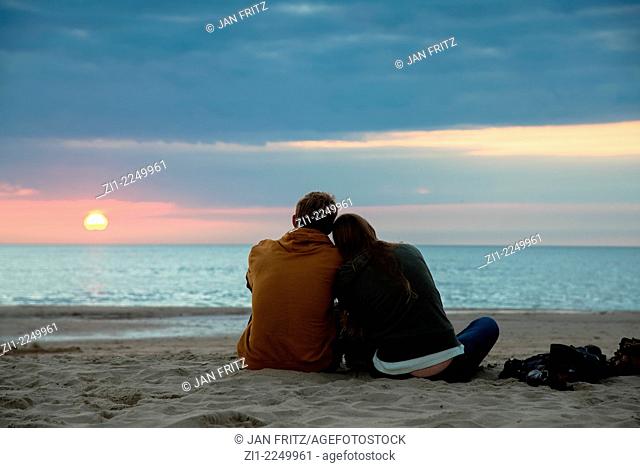 A couple sitting on the sand at the beach enjoying the sunset