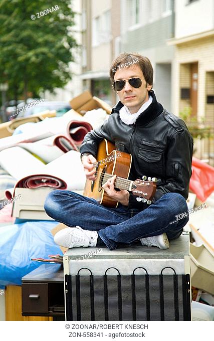 Junger Mann sitzt auf einem alten Kühlschrank vor einem Haufen Sperrmüll und spielt Gitarre