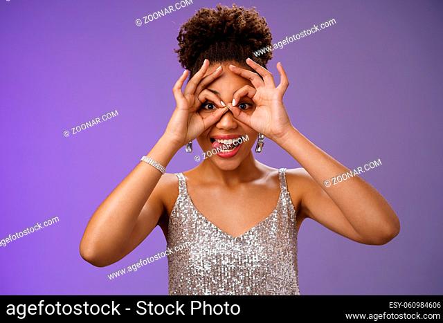 Playful funny good-looking african woman in afro hairstyle in silver glittering dress having fun show tongue smiling broadly make okay ok gesture on eyes look...