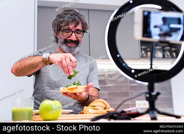 Happy senior vlogger making sandwich with croissant on kitchen island at home