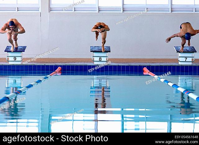 Front view of three male swimmers at swimming pool, jumping from starting blocks, plunging into water. Swimmers training hard for competition