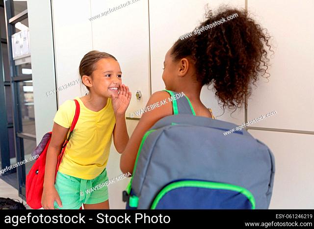 Multiracial elementary schoolgirls gossiping while standing by lockers in school corridor