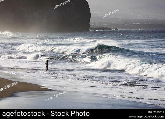 A woman standing on the sandy beach of Praia de Santa Barbara in stormy sea with high waves, Ribeira Grande, Sao Miguel Island, Azores, Portugal, Europe