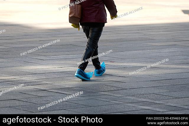23 March 2020, North Rhine-Westphalia, Dortmund: A woman with rubber gloves on her hands and plastic overshoes on her shoes walks through the city centre