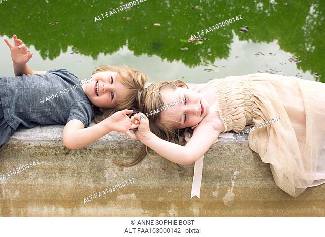 Young sisters lying down on beside pond, holding hands