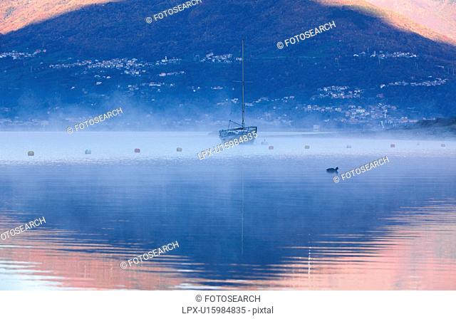 Sailing boat moored at Lake Como, , golden light of sunrise reflected in water with swirling mists, backdrop of mountain slopes, Lombardy, Northern Italy