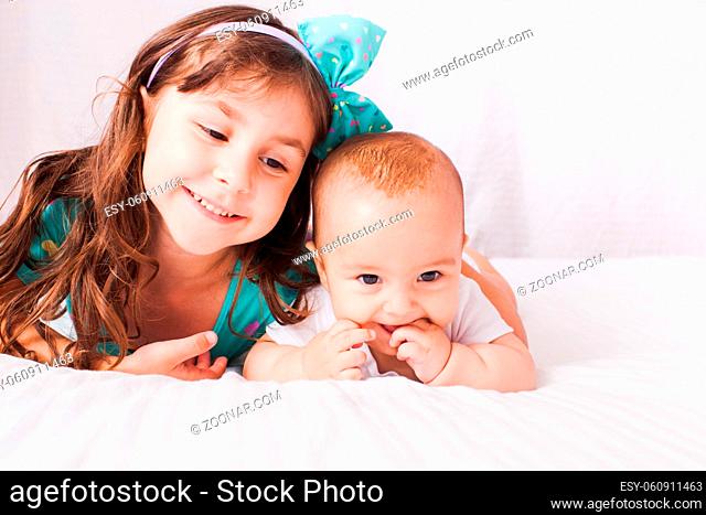The smiling little girl with baby are lying on white sheets in the bedroom