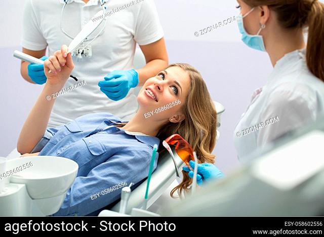 Smiling girl in blue shirt with a mirror in the right hand on the patient chair in the dental cabinet. Next to her there is a dentist and a female assistant