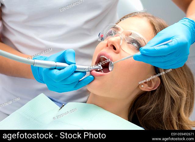 Courageous girl with opened mouth in patient bib and protective eyewear. Next to her there is a dentist in a white uniform with blue latex gloves