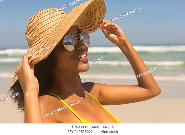 Close-up of young African American woman in yellow bikini, hat and sunglasses standing on beach in the sunshine. She is smiling
