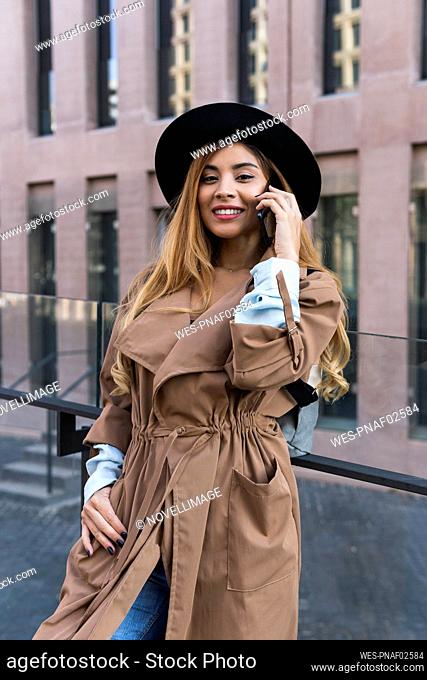 Smiling woman with trenchcoat talking on mobile phone in front of glass railing