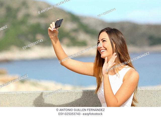 Casual elegant woman greeting with her hand while photographing a selfie with a smartphone on the beach