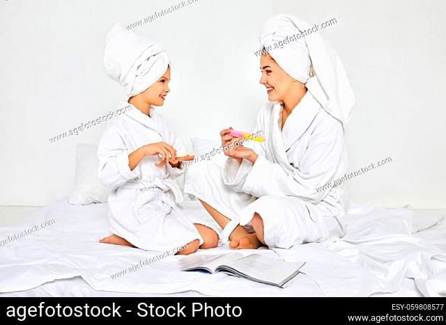 happy mother and her daughter in white bathrobes and towels on heads holding nail file and making manicure