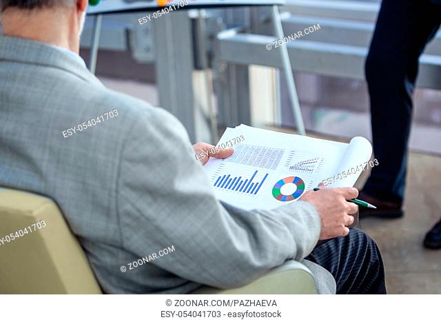 Senior business man sitting at conference, listening to presentation and holding documents with financial diagrams and data