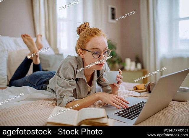 Woman with coffee cup using laptop lying on bed at home